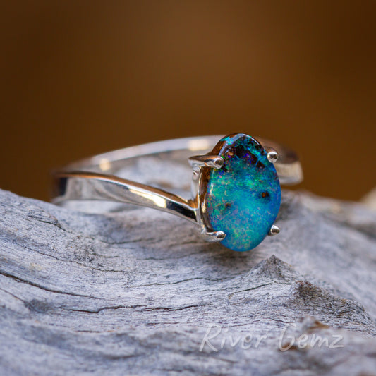 Stunning blue, green ocean like colours evident in the free-form cut opal securely set with four claws in the silver ring. Ring is pictured on a light-grey grained wood and orange/brown backdrop.