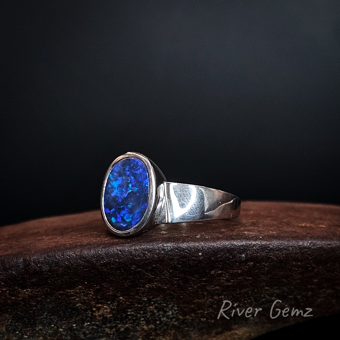Oval shaped blue and purple opal set in silver ring on a brown surface with 'River Gemz' branding.
