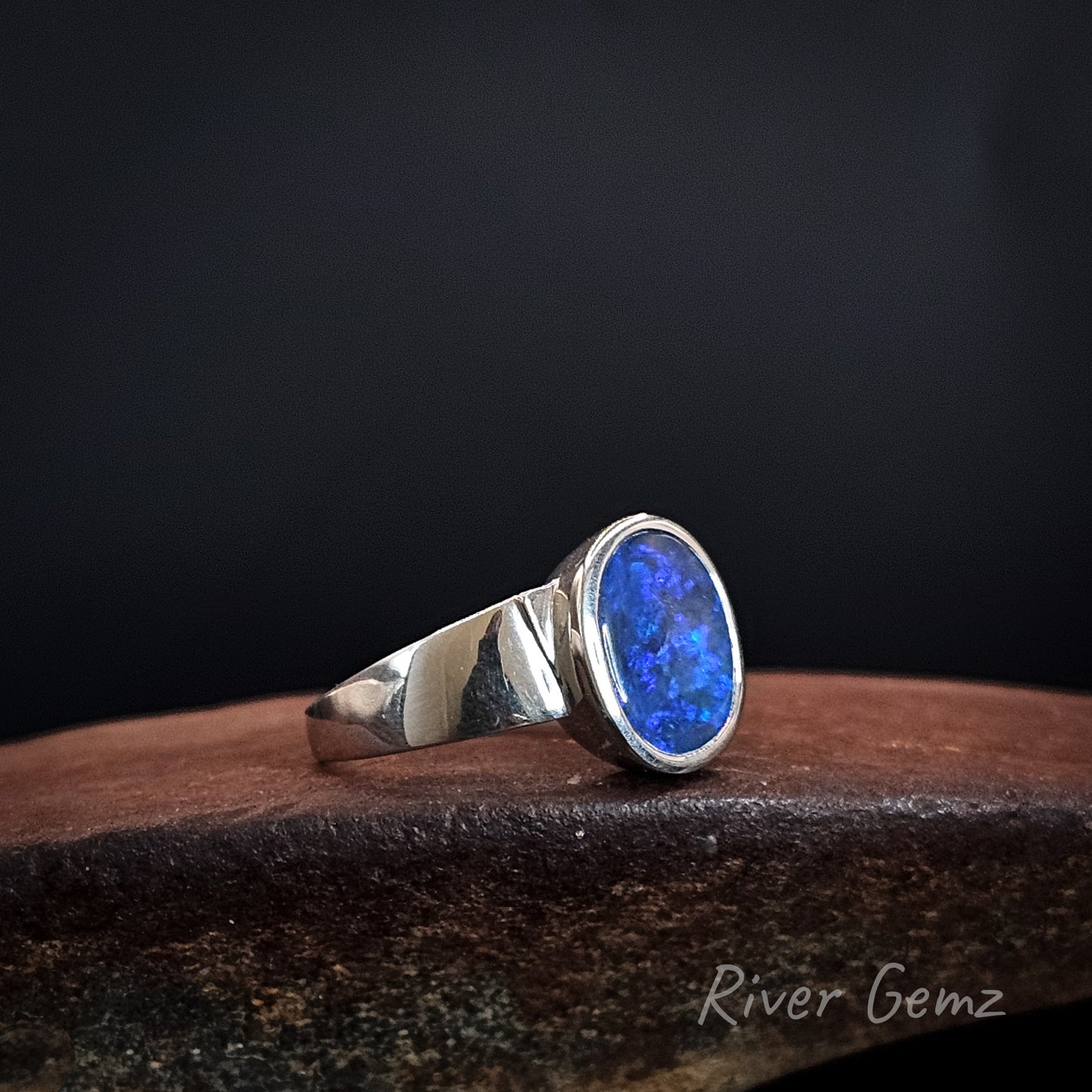 Blue boulder opal set in sterling silver ring with tapered shoulders on a rusty shoe anvil and black background.