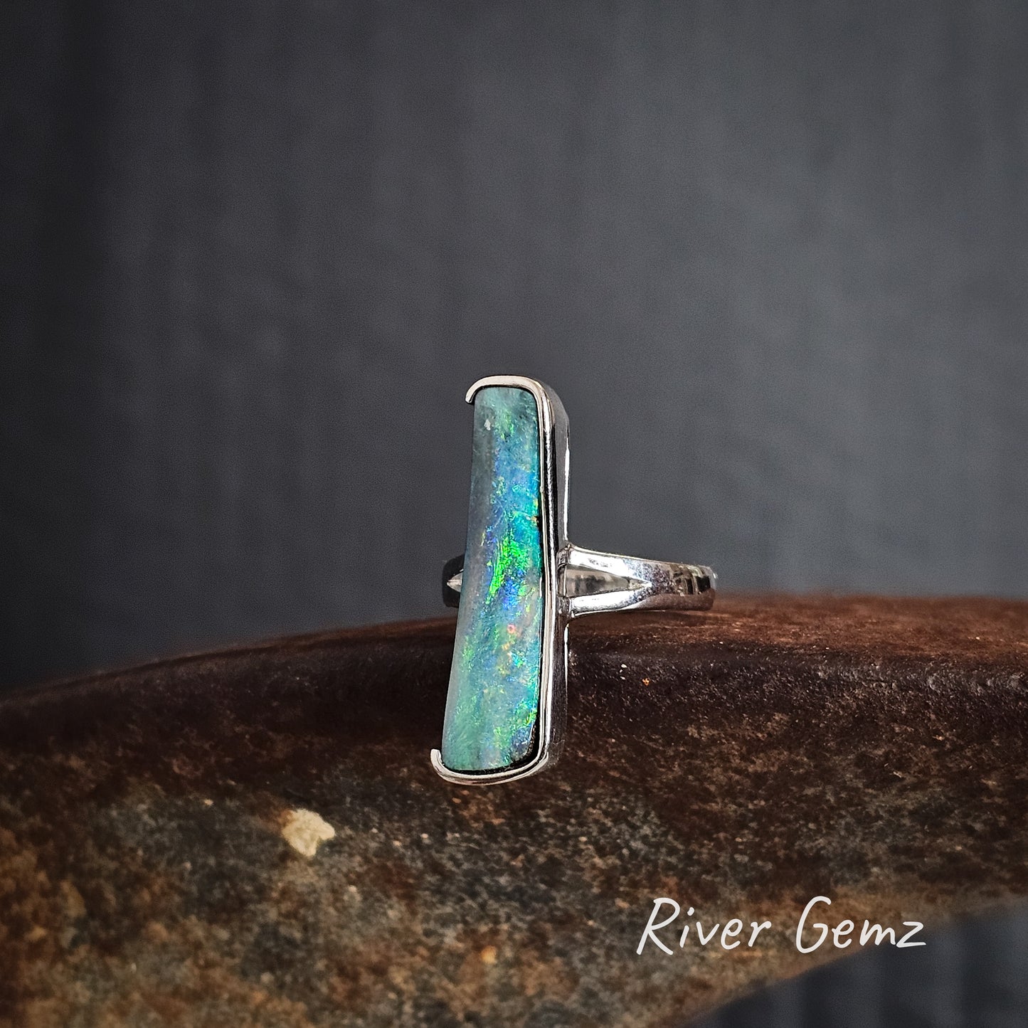 Silver ring with a partial besel set colorful gemstone on a dark surface, branded 'River Gemz'.