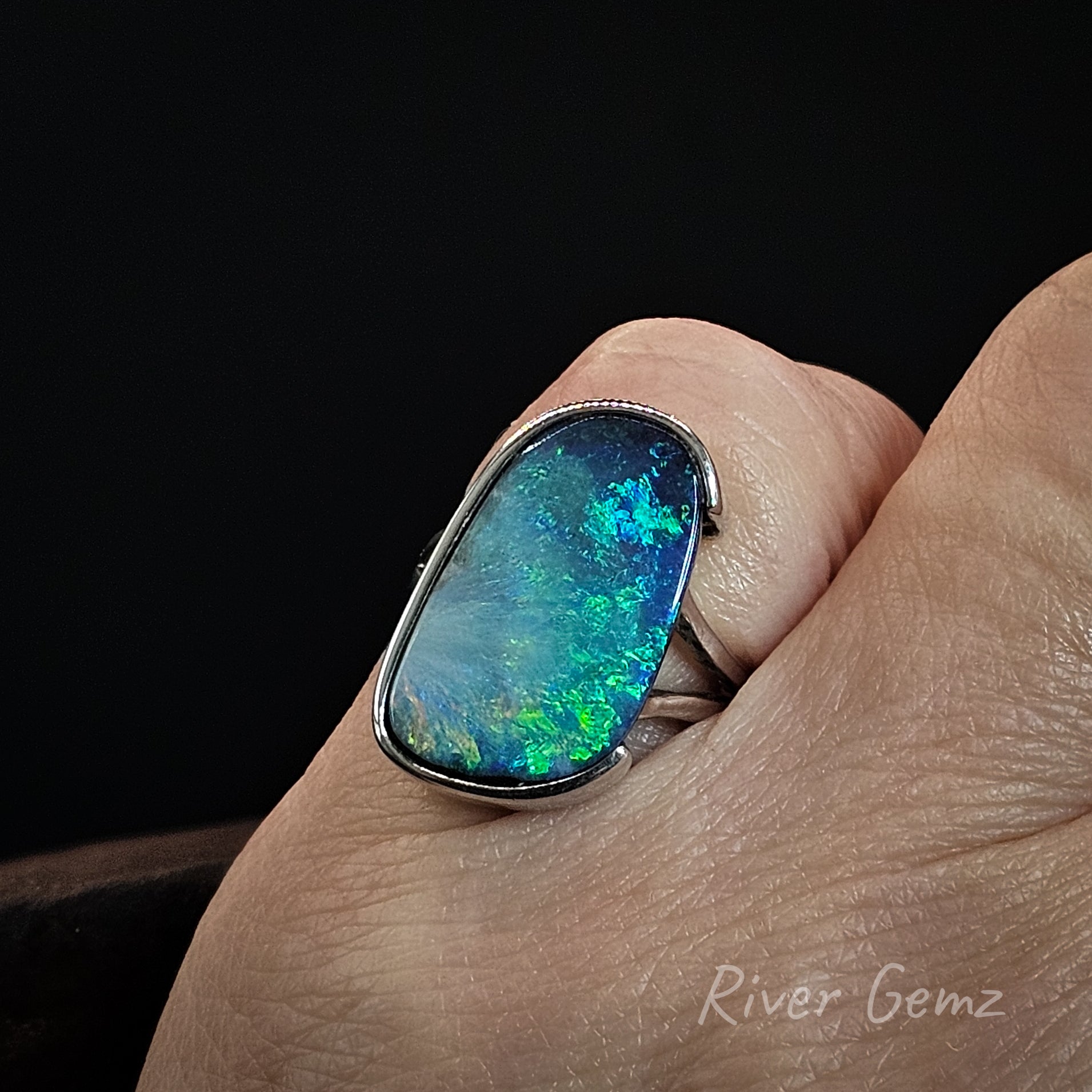 A close-up of a hand wearing a silver ring with a large, irregularly shaped opal gemstone that displays vibrant blue, green, and white play-of-color against a solid black background.