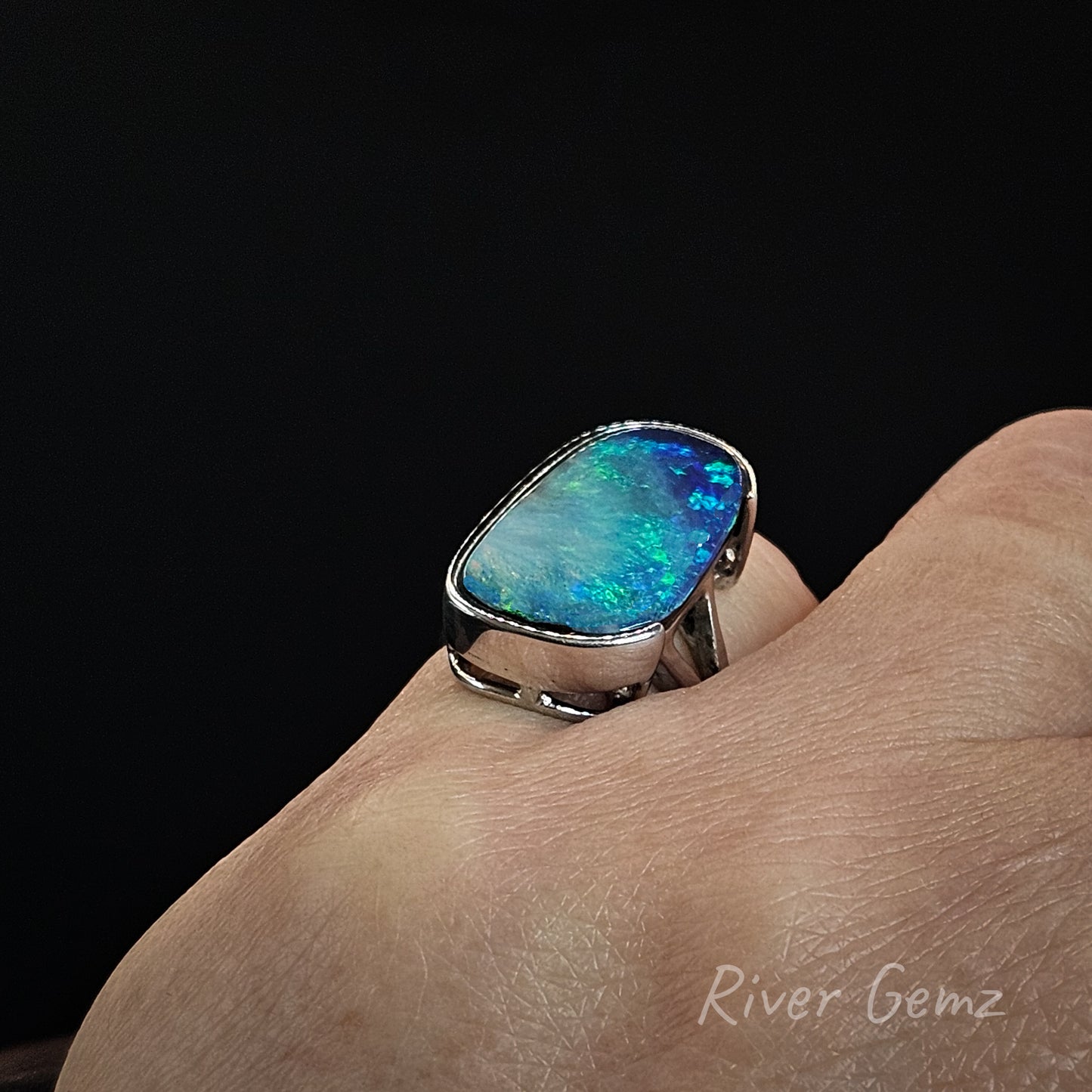 A silver ring with a large, vibrant blue and green opal gemstone, shown worn on a person's finger against a solid black background.