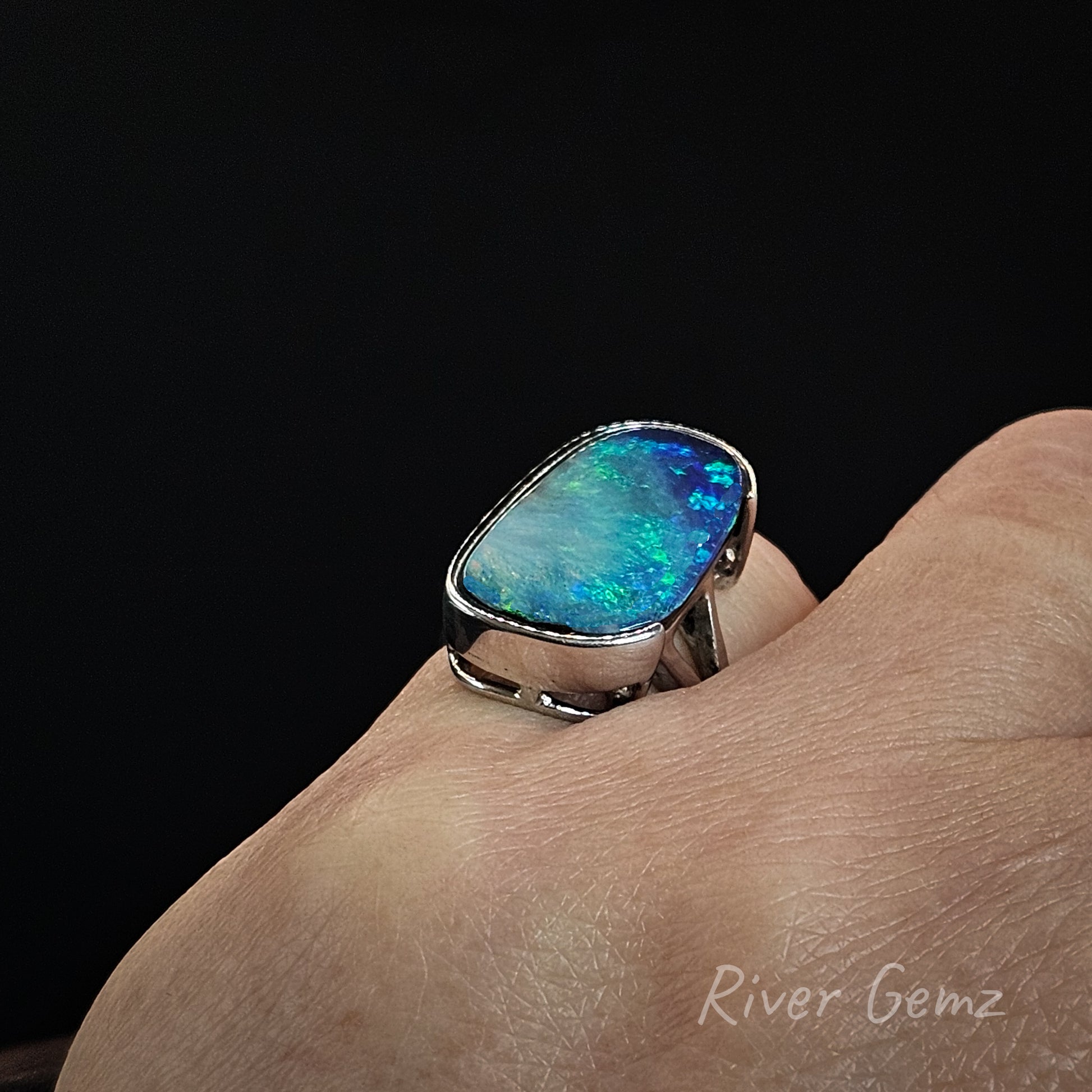 A silver ring with a large, vibrant blue and green opal gemstone, shown worn on a person's finger against a solid black background.