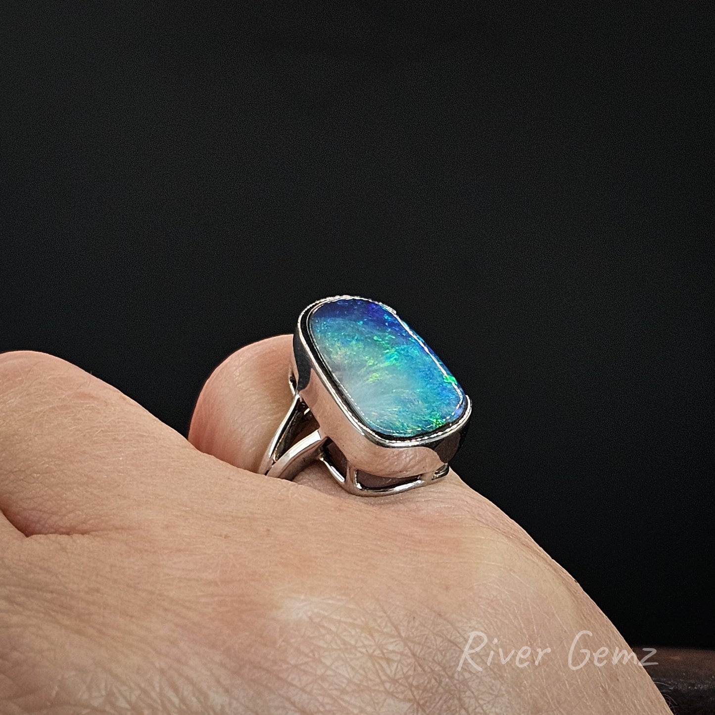 A silver ring with a large, vibrant iridescent opal gemstone displayed on a person's finger against a dark background.