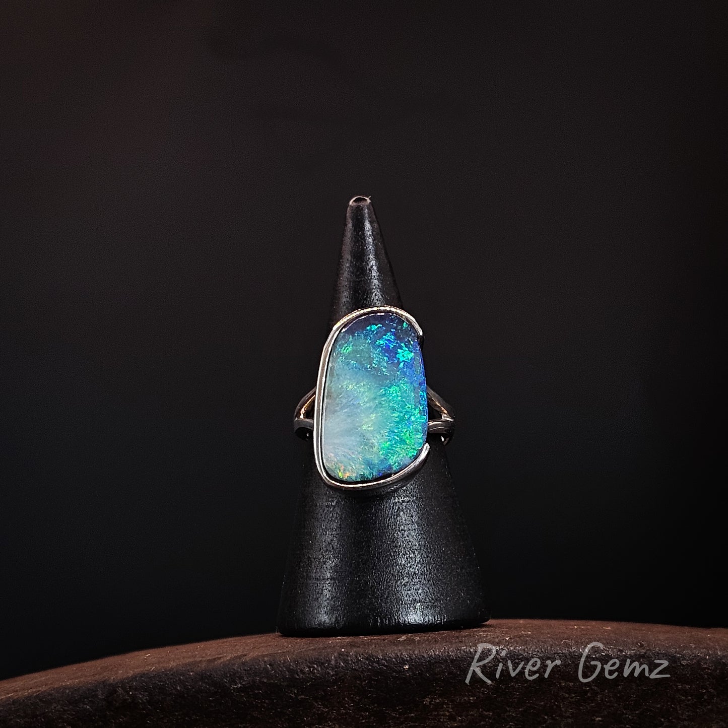 A silver ring with a large, irregularly shaped opal gemstone displaying vibrant blue, green, and white flashes, displayed on a black cone stand against a dark background.