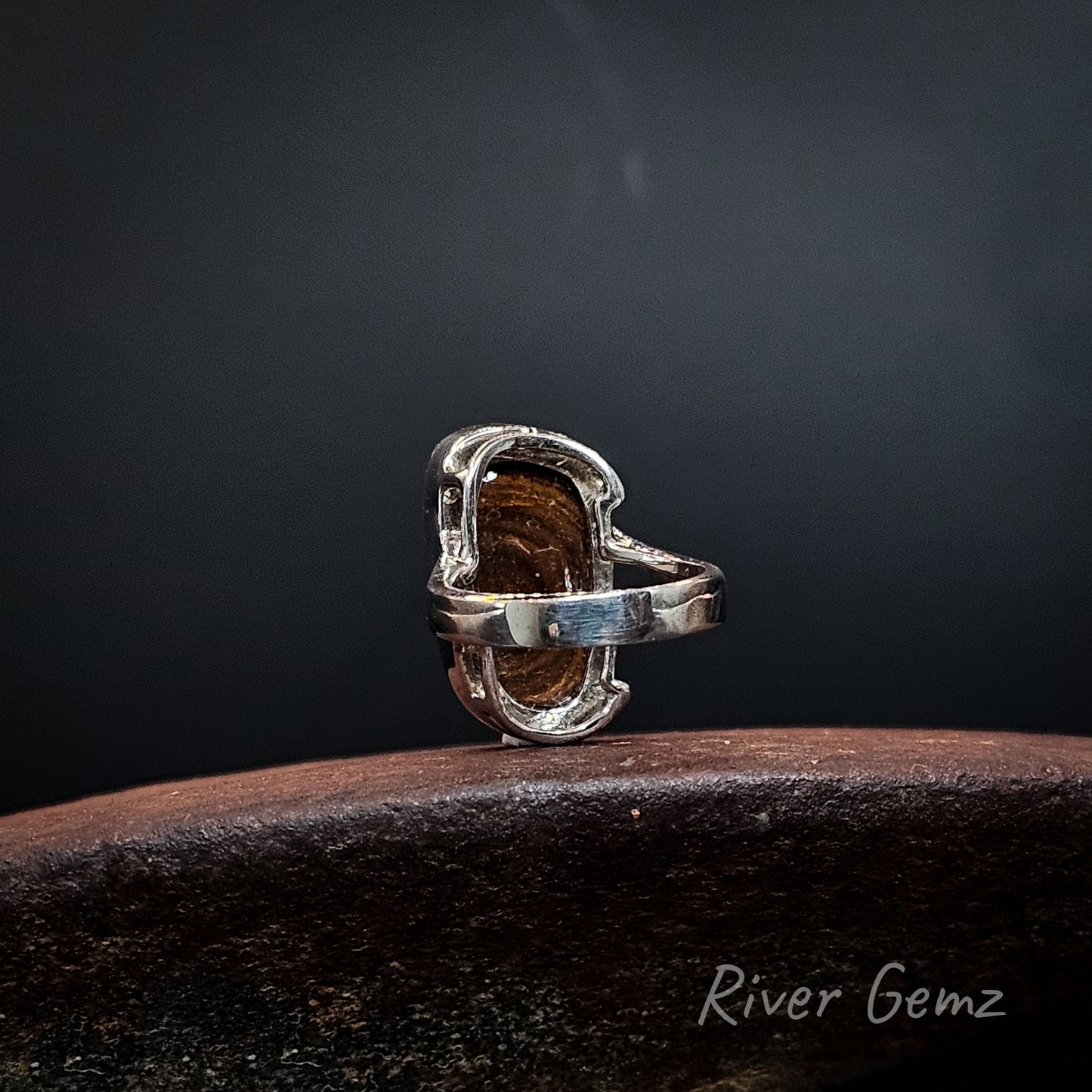 Rear view of a silver ring showing the ironstone matrix base of the boulder opal resting on a dark wooden surface against a dark gray background. The brand name 'River Gemz' is visible in the bottom right corner.