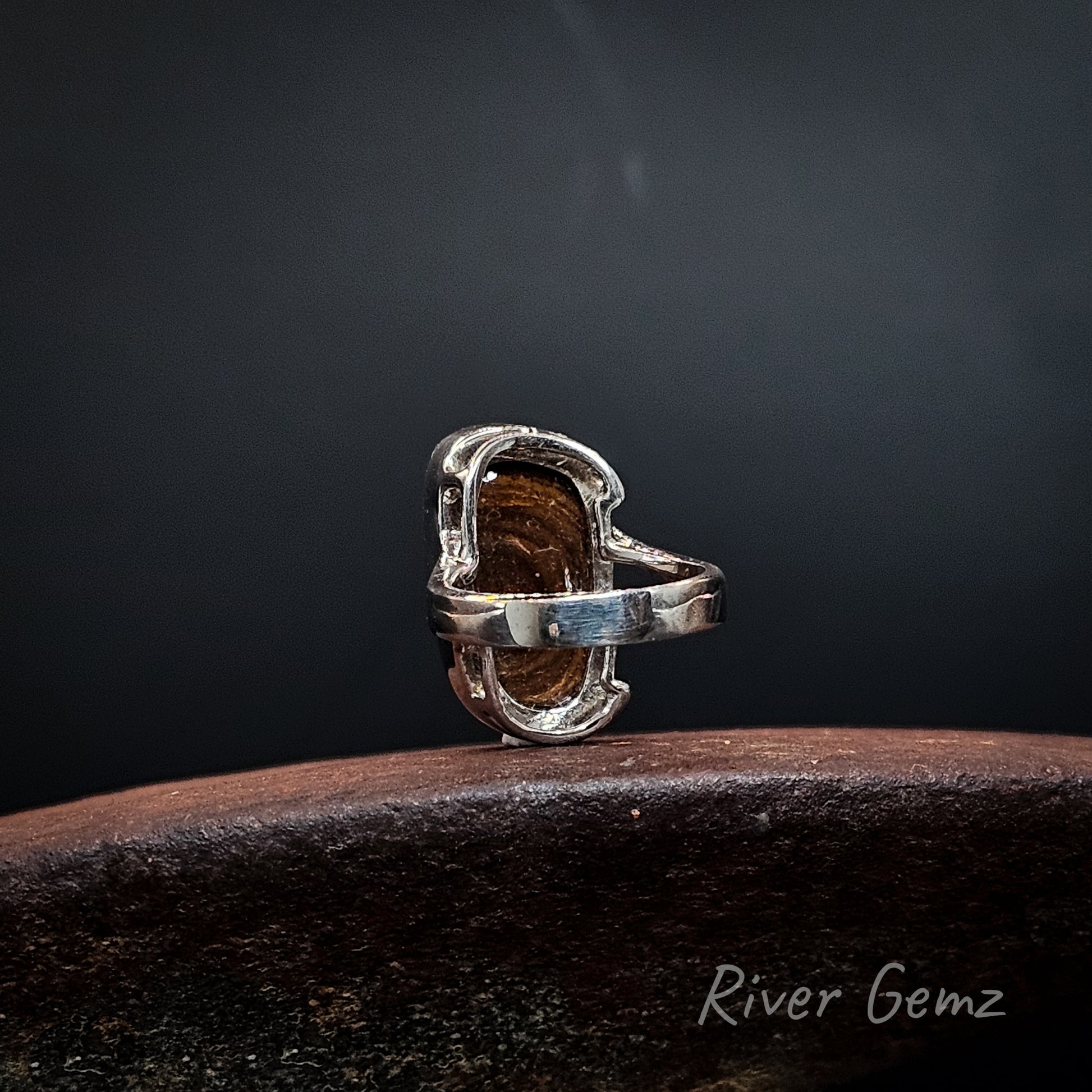 Rear view of a silver ring showing the ironstone matrix base of the boulder opal resting on a dark wooden surface against a dark gray background. The brand name 'River Gemz' is visible in the bottom right corner.