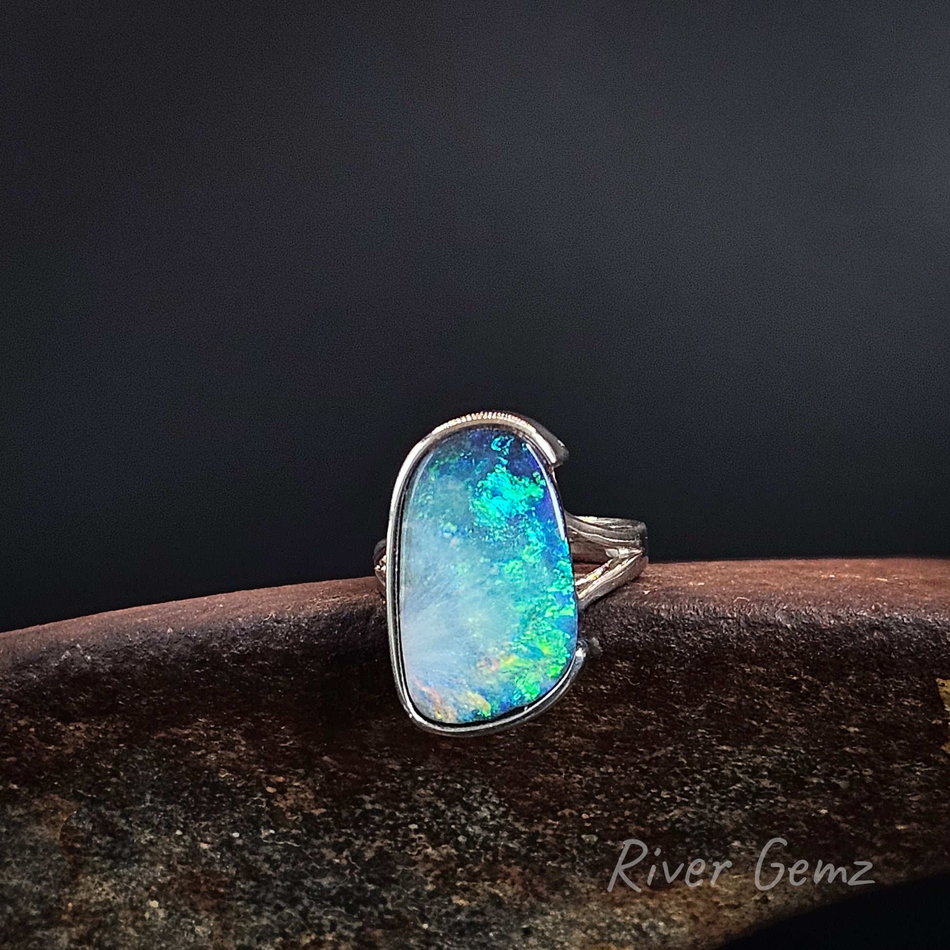 A sterling silver ring featuring a large, irregular-shaped opal gemstone with vibrant blue, green, and white play-of-color, displayed on a dark textured surface against a dark gray background.