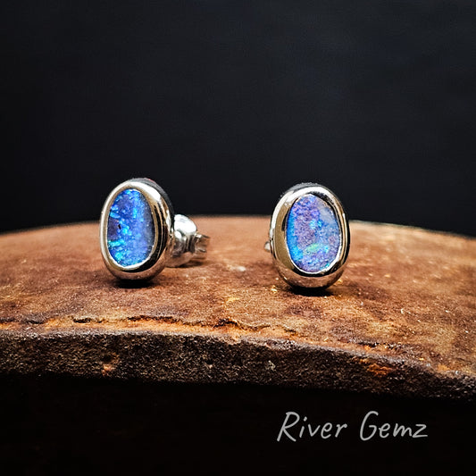 Oval shaped blue & green opals besel set in silver stud earrings photographed on rusty surface & black backdrop.