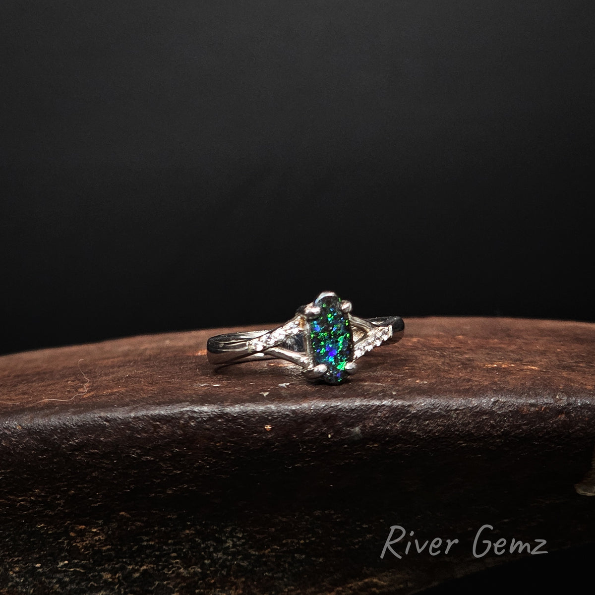 Oval shaped blue and green opal and white topaz claw set in sterling silver ring on a shoe anvil with 'River Gemz' water mark in the bottom right hand corner.