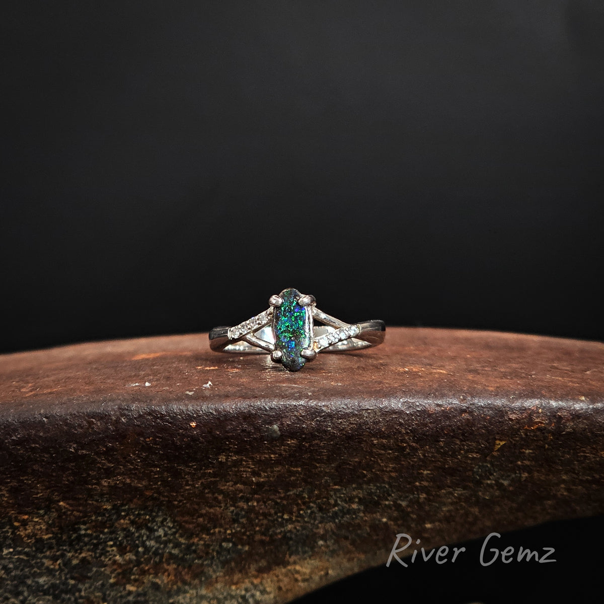 Oval shaped blue and green opal claw set in sterling silver ring on a brown platform and 'River Gemz' branding.