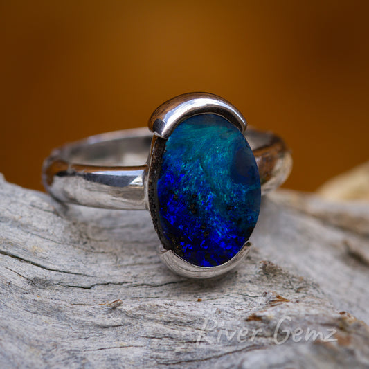 Approximately oval shaped opal is secured above and below with a partial besel setting. The silver ring is seen on a grey weathered piece of wood and a burning orange back ground.