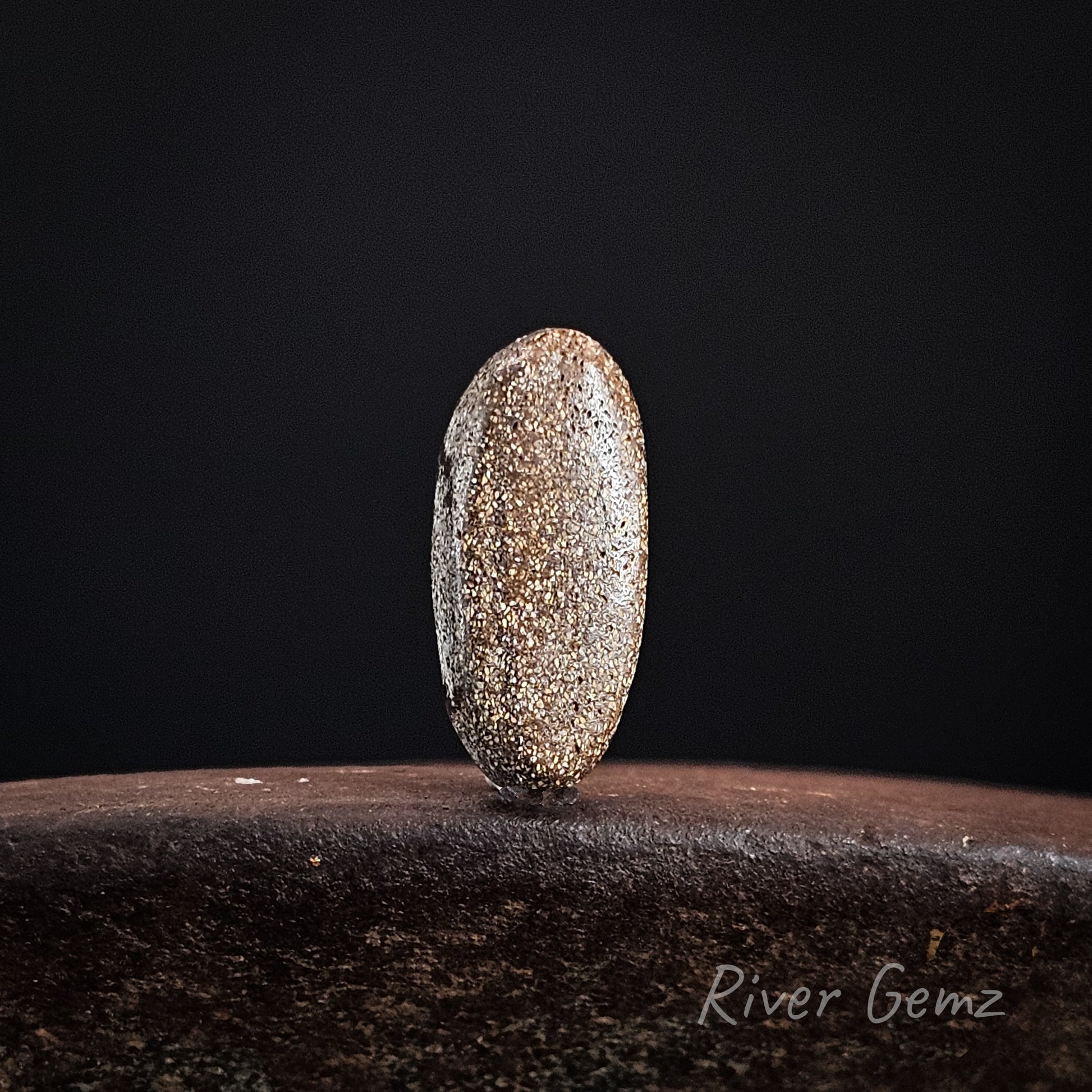 Rear view of oval stone showing the speckled brown surface of the ironstone matrix on a dark background with 'River Gemz' branding.