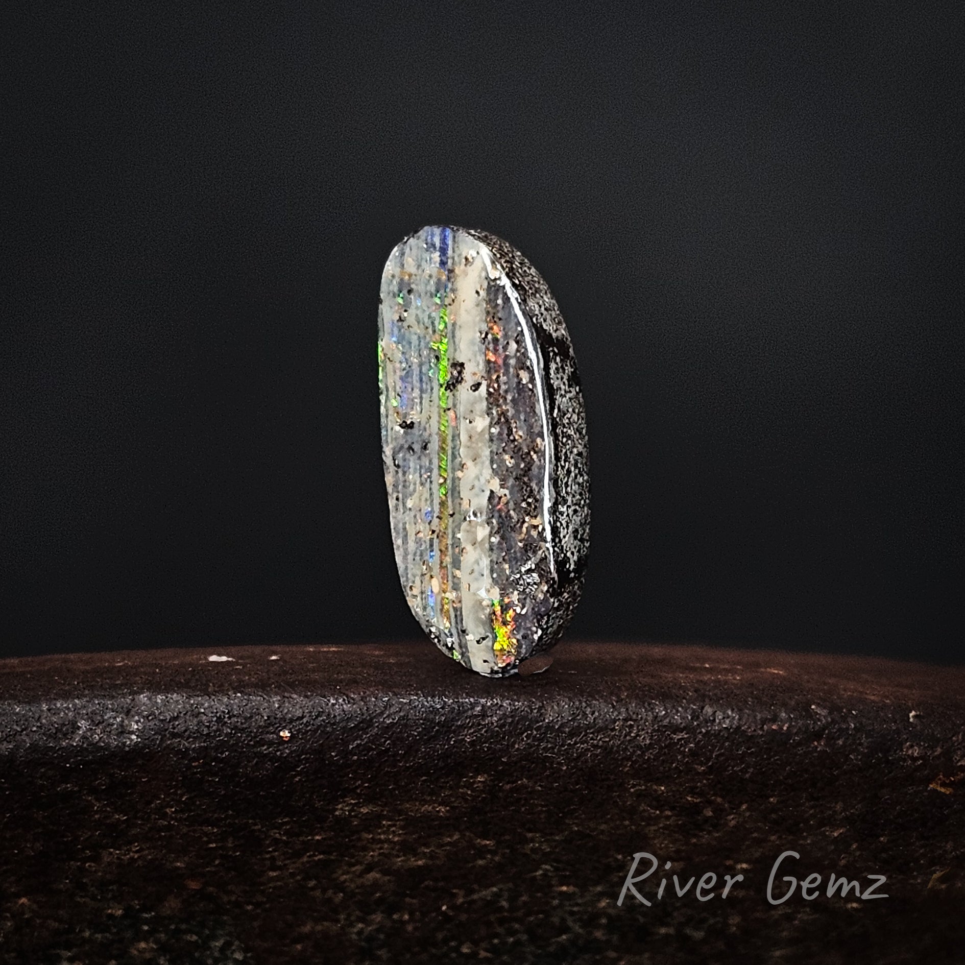 Multicolored stone on a dark surface with 'River Gemz' branding.