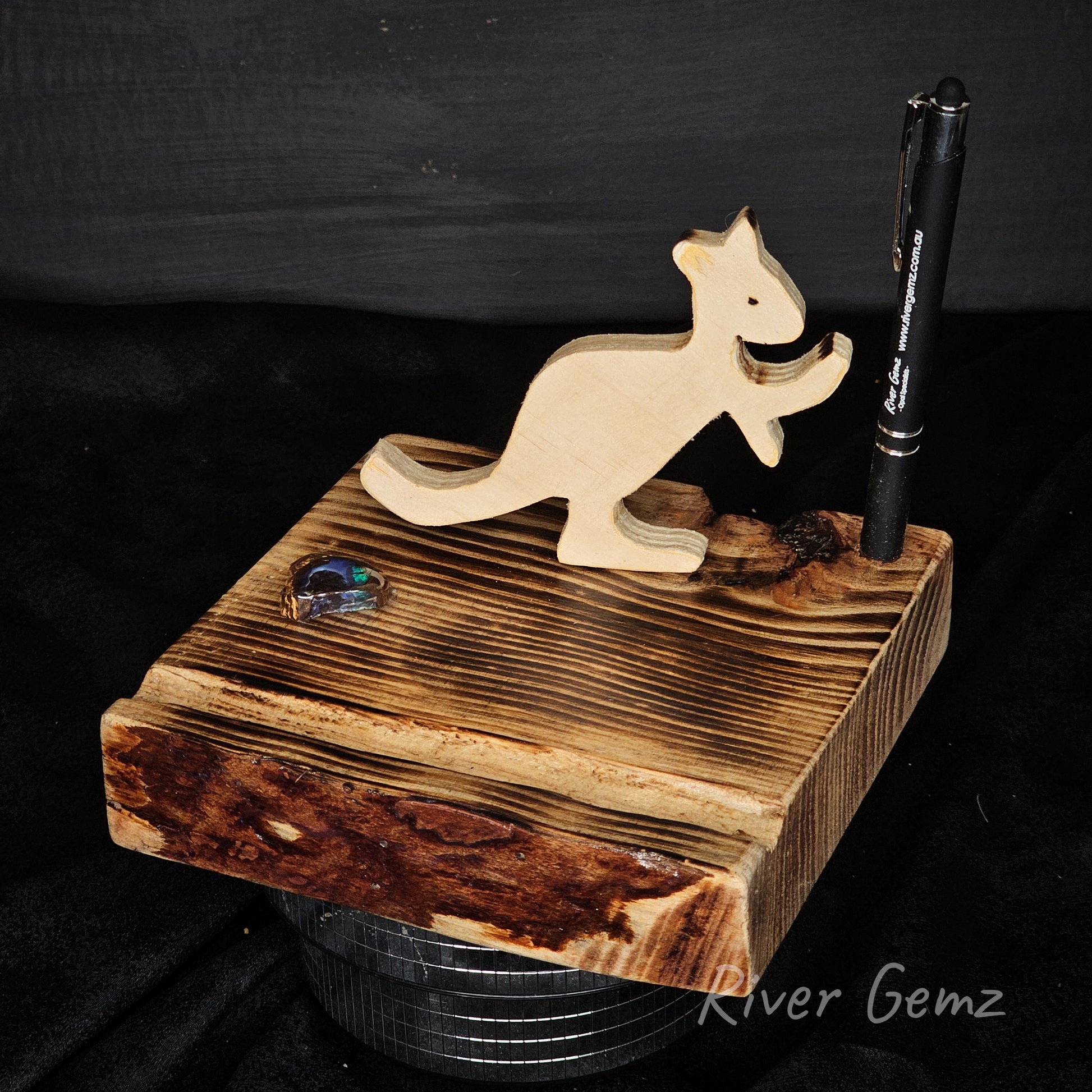 Wooden kangaroo figurine on a wooden stand with a pen and boulder opal specimen, against a dark background.