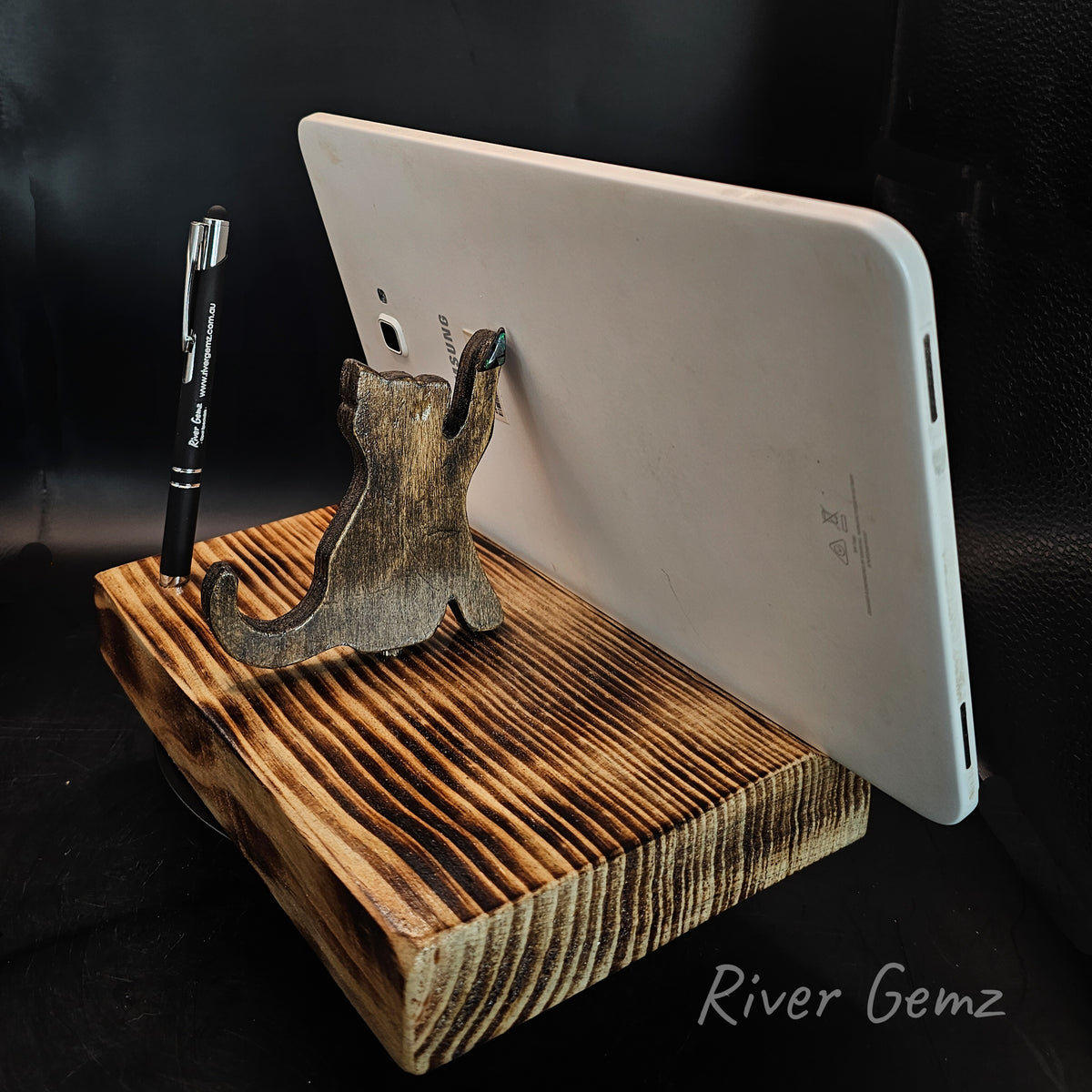 Wooden brown coloured cat is holding a table upright. A pen is upright on the torched wooden base of the phone/tablet stand.