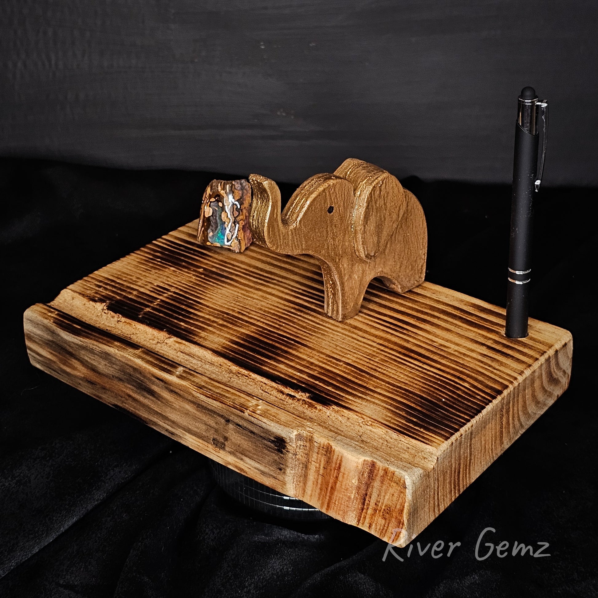 Wooden elephant stand with a pen on a black background, branded 'River Gemz'.