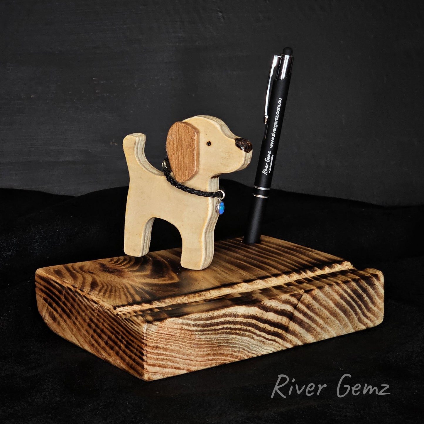 Wooden dog-shaped phone/tablet stand with a stylus on a dark background, branded 'River Gemz'.