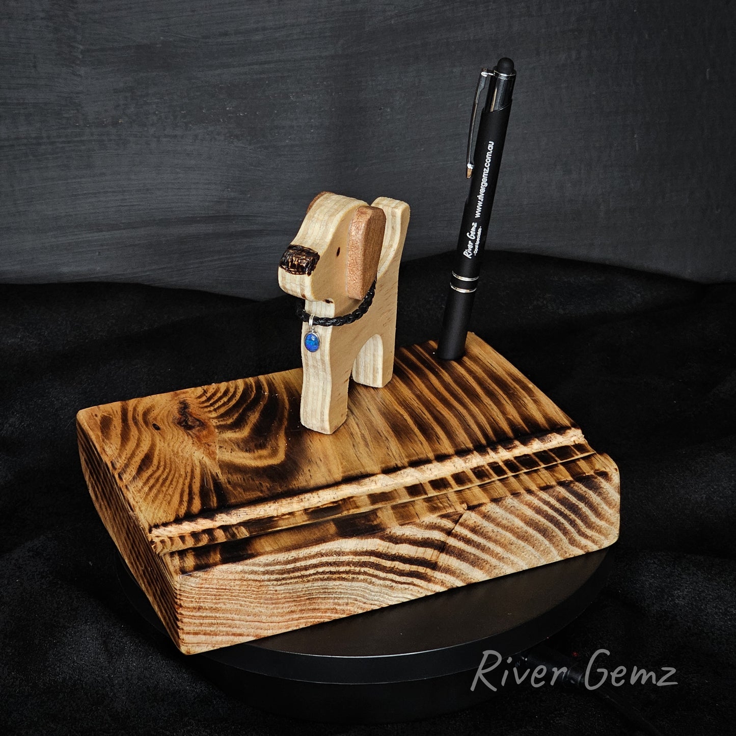 Wooden dog-shaped phone/tablet stand with a pen on a dark background, branded 'River Gemz'.