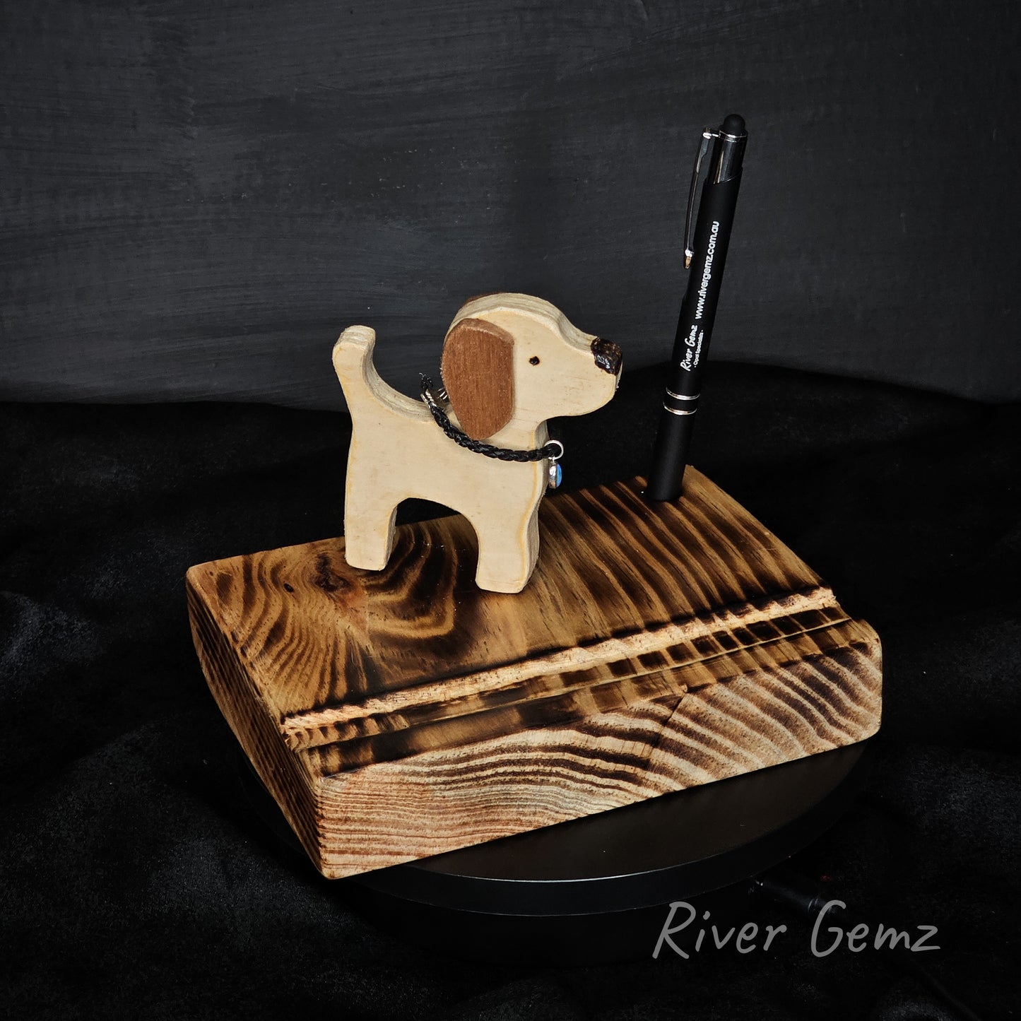 Wooden dog-shaped pen holder with a pen on a dark background, branded 'River Gemz'.